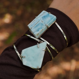 May include: Two silver-toned bracelets with large, light blue rectangular and geometric stones. The stones have a marbled appearance. The bracelets are worn on a brown sleeve, with a blurred natural background.