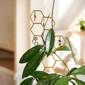 May include: A gold-coloured metal plant trellis in a honeycomb design, decorated with small, colourful beaded charms. The trellis supports a green plant with large, oval leaves, creating a decorative and functional piece for indoor gardening.