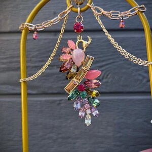Peut inclure: Une pièce décorative dorée avec un motif floral. La pièce présente un groupe de pierres précieuses colorées dans les tons de rose, vert, violet et transparent. La pièce est suspendue à une chaîne dorée et à une arche dorée.