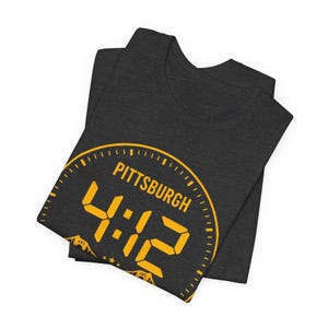 May include: Dark grey t-shirt featuring a yellow clock design. The clock face reads "PITTSBURGH 4:42". The folded shirt displays the design on the front. The design includes a clock face with a digital-style font.