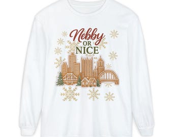 May include: White long-sleeve shirt with a gingerbread-style cityscape graphic. The design includes buildings, bridges, and snowflakes, with the text "Nebby or Nice" in red and green. A festive holiday-themed apparel item.