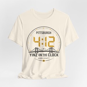 May include: Off-white t-shirt with a gold and black circular design. The design includes the text "PITTSBURGH", the numbers "4:12", a bridge illustration, and the words "YINZ ON THE CLOCK" and "2026 DRAFT".