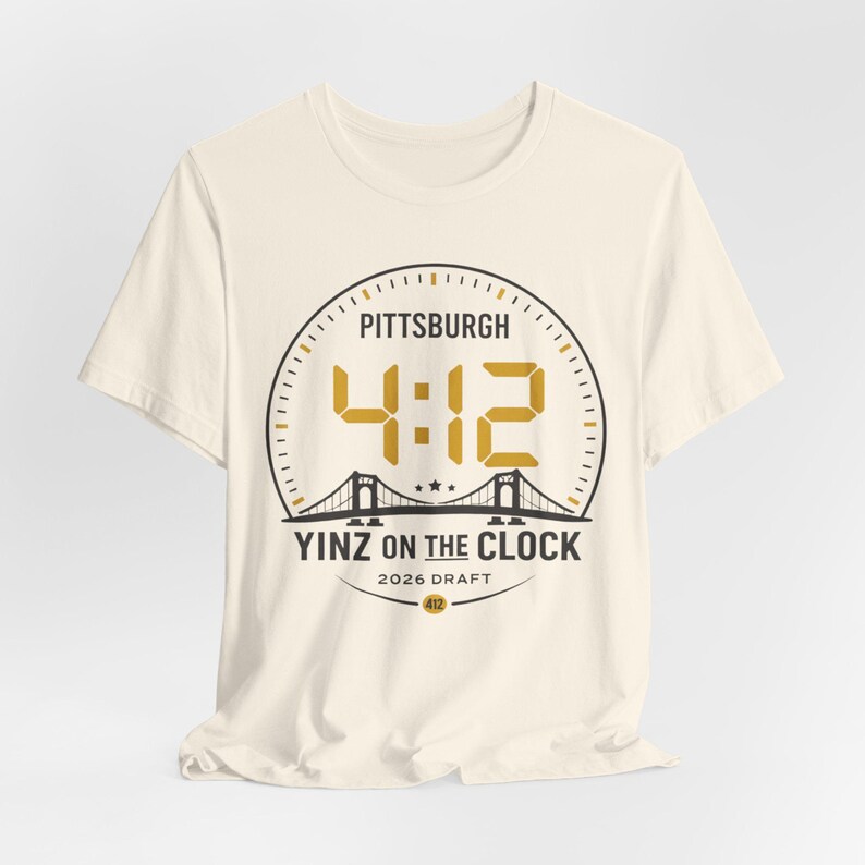 May include: Off-white t-shirt with a gold and black circular design. The design includes the text "PITTSBURGH", the numbers "4:12", a bridge illustration, and the words "YINZ ON THE CLOCK" and "2026 DRAFT".