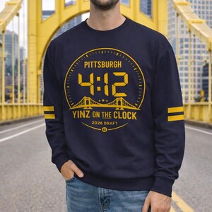Puede incluir: Sudadera azul marino con un gráfico amarillo que dice "PITTSBURGH 4:12 YINZ ON THE CLOCK 2026 DRAFT 412". La sudadera tiene rayas amarillas en las mangas. El fondo presenta un puente amarillo.