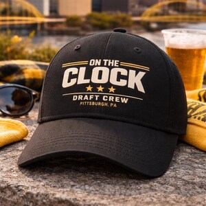 May include: Black baseball cap with the text "ON THE CLOCK DRAFT CREW PITTSBURGH, PA" in white and gold lettering. The cap is on a stone surface with a city skyline in the background.