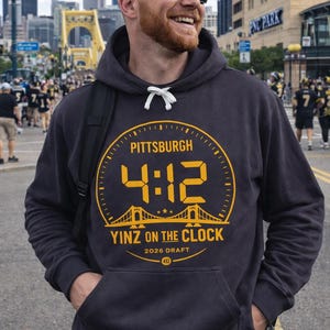 May include: Dark grey hoodie featuring a yellow graphic with the text "PITTSBURGH 4:12 YINZ ON THE CLOCK" and a bridge design. The hoodie has a drawstring hood and a front pocket. The text "2026 Draft 412" is also included.
