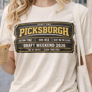 Puede incluir: Camiseta color crema con un diseño gráfico de boleto en negro y dorado. El boleto dice "PICKSBURGH DRAFT WEEKEND 2026". El diseño incluye las frases "ADMITIR UNO", "SECCIÓN: YINZ", "FILA: 412" y "ASIENTO: EN EL RELOJ".