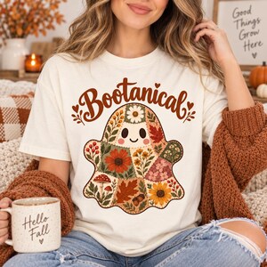 May include: A cream-coloured t-shirt featuring a graphic of a ghost filled with a patchwork pattern of autumn flowers, leaves, mushrooms, and acorns. The text above the ghost reads Bootanical in a brown script font with small heart accents.