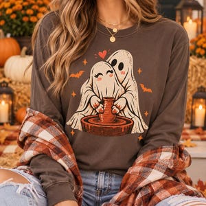 Könnte beinhalten: Ein dunkelgraues Langarmshirt mit einer Grafik von zwei weißen Geistern, die gemeinsam an einer Töpferscheibe arbeiten. Das Design enthält orangefarbene Fledermäuse, kleine Sterne und ein rotes Herz über den Geistern.