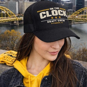 May include: Black trucker hat featuring the text "ON THE CLOCK" in white and yellow, with "DRAFT CREW PITTSBURGH, PA" underneath. The hat is worn by a person in a yellow hoodie and denim jacket, with a city skyline in the background.