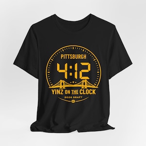 May include: Black t-shirt with a gold graphic. The graphic features the word "Pittsburgh" above the numbers "4:12" in a clock-like design. Below the numbers is "Yinz on the Clock" and "2026 Draft".