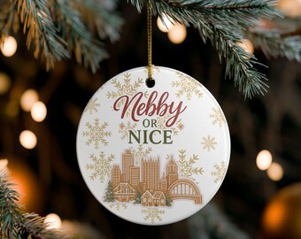 May include: A white ceramic Christmas ornament with the words "Nebby or Nice" in burgundy and green. The ornament features a gingerbread-style cityscape with gold snowflakes and a gold hanging string.