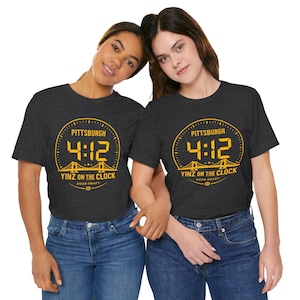 May include: Two people wearing dark grey t-shirts featuring a yellow graphic. The graphic includes the word "Pittsburgh", the numbers "4:12", a bridge illustration, and the text "Yinz on the Clock 2026 Draft". They are wearing blue jeans.