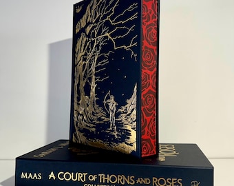 Puede incluir: Un libro de edición de coleccionista titulado "A Court of Thorns and Roses" de Maas. La portada azul marino presenta ilustraciones doradas de una escena de bosque. El lomo es rojo con un patrón de rosas.