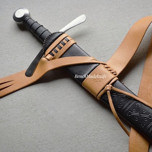 May include: A medieval sword in a black scabbard with a tan leather strap and accents. The sword has a silver pommel and a black handle. The scabbard is decorated with a geometric pattern. The image includes the text "Bench Madeknife".
