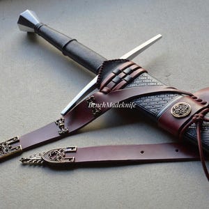 May include: A detailed image of a sword with a silver blade and a black handle. The sword is sheathed in a brown leather scabbard with decorative metal accents. The scabbard has a matching belt. The text "BenchMadeKnife" is visible.