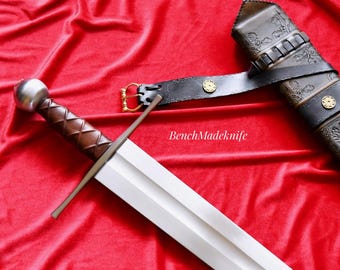 May include: A medieval sword with a silver pommel and a brown leather-wrapped handle, displayed on a red velvet surface. The sword has a long, straight, double-edged blade. A black leather sheath with decorative tooling and a belt are also present.