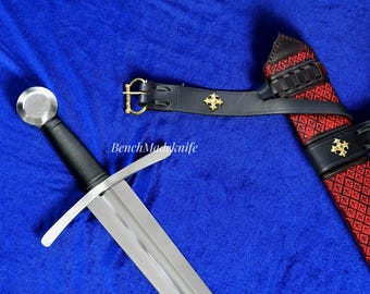 May include: A medieval sword with a silver blade and a black handle, set against a blue backdrop. The sword is displayed with a black leather scabbard featuring red diamond-patterned fabric and gold detailing. The scabbard has a black leather belt with a gold buckle.
