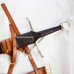 Può includere: Una spada in stile medievale con una lama d'argento e un'impugnatura scura con motivi intricati. La spada è in un fodero di cuoio marrone, fissato da cinghie di cuoio marroni e fibbie color oro. Il testo "BenchMadeKnife" è visibile.
