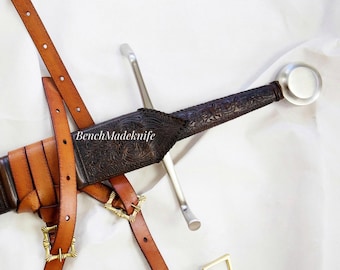 May include: A medieval sword with a silver blade and a dark, patterned handle. The sword is in a brown leather sheath, secured by brown leather straps and gold-coloured buckles. The text "BenchMadeKnife" is visible on the sheath.