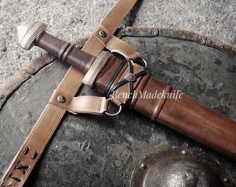 May include: A medieval sword with a brown leather scabbard and tan leather straps, lying on a dark, weathered shield. The sword has a silver hilt and a wooden handle. The image has a rustic, historical aesthetic.