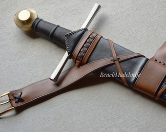 May include: A medieval-style sword with a black handle and a silver blade, sheathed in a black scabbard with a brown leather belt. The scabbard has an embossed design. The belt has a gold buckle.