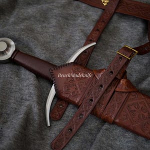 May include: A medieval-style sword with a brown leather handle and sheath. The sheath and belt feature intricate embossed patterns. The sword's blade is silver, and the handle has a silver pommel. The text "BenchMadeknife" is visible on the sheath.