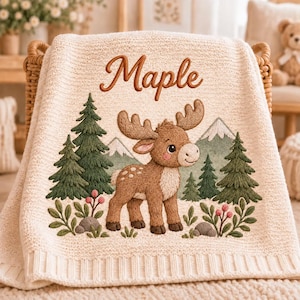 May include: A cream-coloured knit blanket featuring an embroidered scene of a brown moose standing in a forest with green pine trees, light green mountains, and small pink berries. The name Maple is embroidered in brown script at the top.