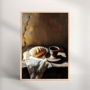 Op de afbeelding: Een ingelijste kunstdruk van een stilleven. Het toont een brood op een witte doek, een donkerbruine beker en een schotel op een rustieke houten tafel. De achtergrond is een getextureerde, sepia-getinte muur met zonlicht.