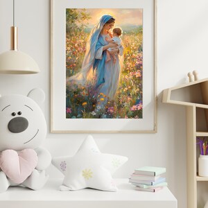 May include: A framed print depicts a woman in a blue robe holding a baby in a field of flowers. The artwork has a soft, painterly style with warm lighting. The frame is a light wood color, and the print is displayed in a room with a white shelf and decorative items.