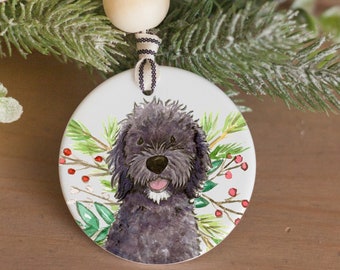May include: A round ceramic Christmas ornament featuring a watercolor illustration of a black dog with a happy expression. The ornament is decorated with greenery and red berries, and hangs from a striped ribbon.