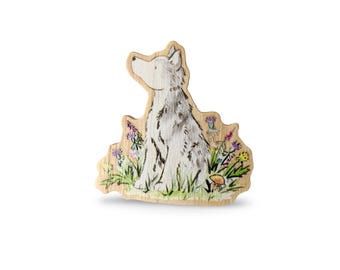 May include: A wooden cutout of a white wolf sitting in a field of flowers. The wolf is looking to the left and has a light gray coat. The flowers are in shades of pink, purple, yellow, and blue. There is a small mushroom in the lower right corner.