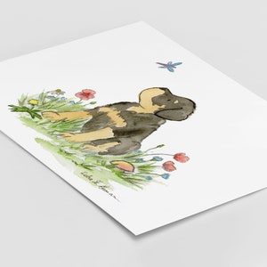 May include: Watercolor painting of a black and brown puppy sitting in a field of flowers. The puppy is looking to the right and there is a dragonfly flying above it. The painting is signed "Lida S. Hamar".