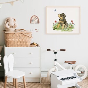 May include: A watercolor illustration of a black and brown puppy sitting in a field of flowers, looking up at a butterfly. The illustration is framed and hanging on a white wall in a nursery.