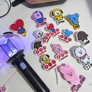 May include: A black light stick with a clear globe containing a purple BTS logo, surrounded by various colorful rubber charms featuring cartoon characters, some labeled with names like J-Hope, Jimin, RM, Suga, and Jin, and others with Arirang text.