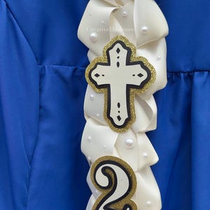 K&ouml;nnte beinhalten: Ein wei&szlig;es Band mit einem Kreuz und der Zahl 2, verziert mit Perlen. Das Kreuz hat einen goldglitzernden Umriss und einen schwarzen Rand. Die Zahl 2 hat ebenfalls einen goldglitzernden Umriss und einen schwarzen Rand. Der Hintergrund ist ein blauer Stoff.