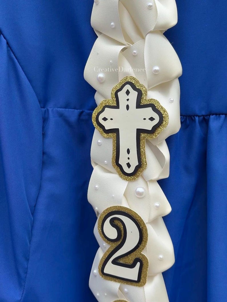 K&ouml;nnte beinhalten: Ein wei&szlig;es Band mit einem Kreuz und der Zahl 2, verziert mit Perlen. Das Kreuz hat einen goldglitzernden Umriss und einen schwarzen Rand. Die Zahl 2 hat ebenfalls einen goldglitzernden Umriss und einen schwarzen Rand. Der Hintergrund ist ein blauer Stoff.