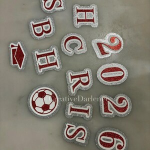 K&ouml;nnte beinhalten: Eine Sammlung von rot-silbernen, glitzernden, ausgestanzten Buchstaben, Zahlen und einem Fu&szlig;ball. Die Buchstaben und Zahlen haben einen wei&szlig;en Rand und sind auf einer hellgrauen Oberfl&auml;che verstreut. Der Fu&szlig;ball ist rot und wei&szlig;.
