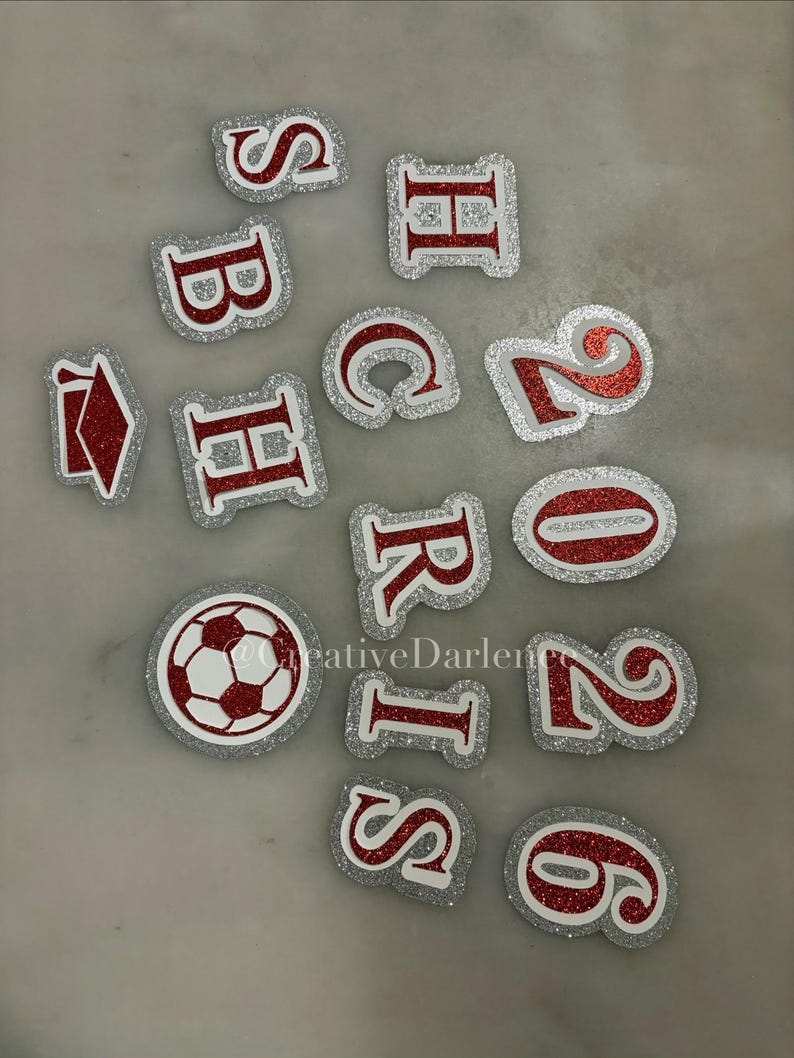 K&ouml;nnte beinhalten: Eine Sammlung von rot-silbernen, glitzernden, ausgestanzten Buchstaben, Zahlen und einem Fu&szlig;ball. Die Buchstaben und Zahlen haben einen wei&szlig;en Rand und sind auf einer hellgrauen Oberfl&auml;che verstreut. Der Fu&szlig;ball ist rot und wei&szlig;.