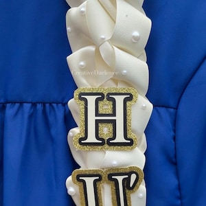 K&ouml;nnte beinhalten: Ein cremefarbenes Band-Lei mit den Buchstaben "H" und "U" in goldfarbenem Glitzer und schwarzem Rand. Das Lei ist mit kleinen, wei&szlig;en Perlen verziert und vor blauem Hintergrund platziert. Der Text "CreativeDarlence" ist sichtbar.