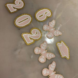K&ouml;nnte beinhalten: Rosa- und goldfarbene Glitzer-Stanzformen, darunter die Zahlen 2026 und mehrere Schmetterlings- und Diamantenformen. Die Formen haben einen goldfarbenen Glitzerrahmen und liegen auf einer hellen Oberfl&auml;che.