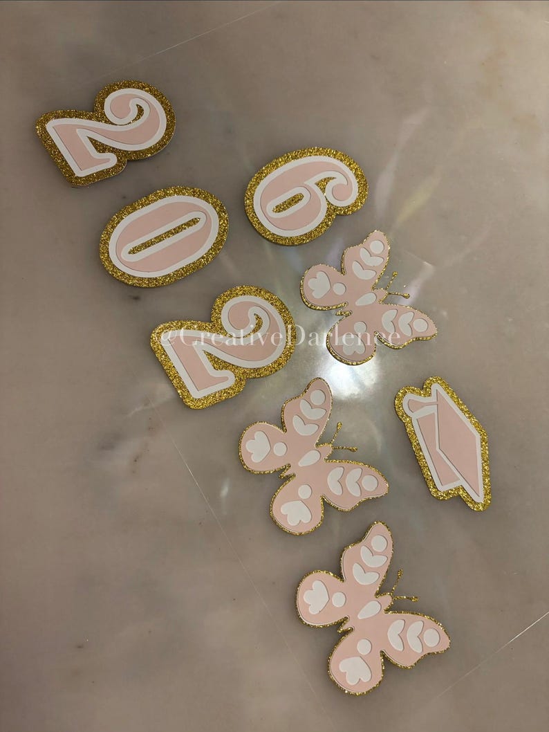 K&ouml;nnte beinhalten: Rosa- und goldfarbene Glitzer-Stanzformen, darunter die Zahlen 2026 und mehrere Schmetterlings- und Diamantenformen. Die Formen haben einen goldfarbenen Glitzerrahmen und liegen auf einer hellen Oberfl&auml;che.