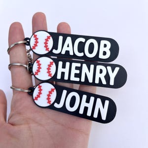 Puede incluir: Tres llaveros negros con texto blanco "JACOB", "HENRY" y "JOHN". Cada llavero tiene un diseño de béisbol con costuras rojas. Los llaveros se sostienen en una mano.