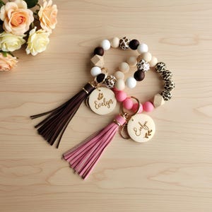 May include: Two beaded wristlet keychains with wooden details and tassels. One has a brown tassel and a wooden charm with a butterfly and the name "Evelyn". The other has a pink tassel and a wooden charm with the name "Evelyn" and a leaf design.