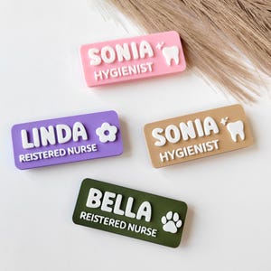 Puede incluir: Cuatro insignias rectangulares con nombres en rosa, morado, beige y verde. Las insignias tienen letras blancas en relieve con nombres y títulos. Dos insignias dicen "SONIA" con "HIGIENISTA" y un gráfico de diente. Una dice "LINDA" con "ENFERMERA REGISTRADA" y una flor. La última dice "BELLA" con "ENFERMERA REGISTRADA" y una huella de pata.
