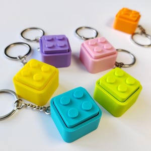 May include: A collection of six square keychains, each featuring a raised four-stud brick design on top. The keychains are coloured in bright yellow, purple, pink, orange, lime green, and turquoise, and each includes a silver metal split ring.