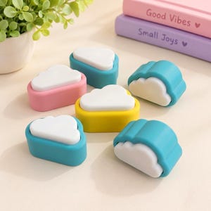 May include: A collection of six cloud-shaped erasers, each featuring a white cloud top set into a colourful base. The bases are coloured in shades of pink, yellow, and various tones of blue. These small, functional stationery items are arranged together.