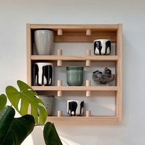 Può includere: Una mensola in legno a parete con più livelli, che espone varie tazze in ceramica e una piccola ciotola di vetro. Le tazze presentano una gamma di colori e motivi, tra cui disegni in bianco e nero e un verde tenue. La mensola è montata su un muro bianco.