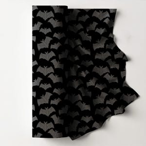 May include: A roll of black fabric featuring a repeating pattern of grey bats. The bats are evenly spaced, creating a Halloween or gothic theme. The fabric is likely for crafting or clothing.