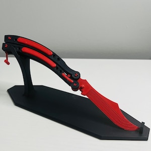May include: A red and black butterfly knife, displayed on a black stand. The blade is red with a ribbed design, and the handle is black with red accents. The stand is a simple, angular design.